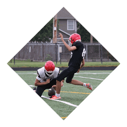 Two people wearing red helmets and sports uniforms are on a football field with one person holding the ball on the ground while the other is kicking it. Description generated by AI