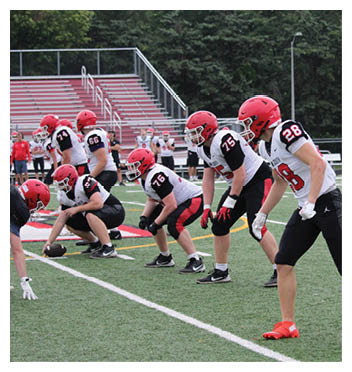 Several people wearing red helmets and football uniforms are positioned on a football field preparing for a play. Description generated by AI