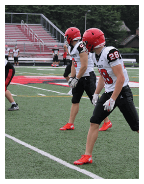 People wearing sports uniforms and cleats are standing on a football field near the 20-yard line. Description generated by AI