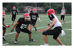 People wearing sports uniforms are engaged in a football game on a grassy field with one person holding a football and others in defensive stances. Description generated by AI