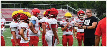 A group of people wearing red and white football uniforms stand on a green field while two others in casual athletic clothing appear to be coaching or instructing them. Description generated by AI