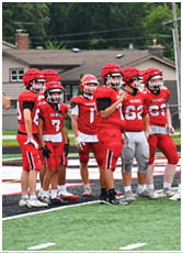 Four people wearing red football uniforms with helmets stand on a field with artificial turf. Description generated by AI