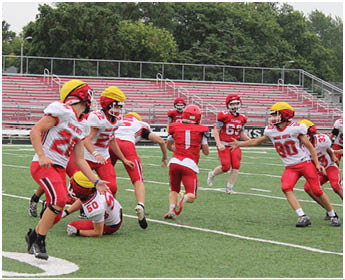 People wearing red and white football uniforms are actively playing on a green field with bleachers in the background. Description generated by AI