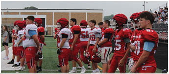 A group of people wearing red and white football uniforms stand in a line on the sidelines of a football field with spectators in the background. Description generated by AI