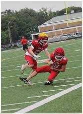 Two people in red football uniforms with helmets are playing on a field, with one person holding the ball while the other runs behind. Description generated by AI