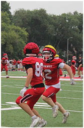 People wearing red sports uniforms are gathered on the sidelines of a green football field with trees and houses in the background. Description generated by AI