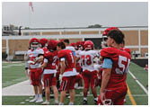 A group of people wearing red and white football uniforms stand on a green field near the sideline with an American flag and scoreboard visible in the background. Description generated by AI