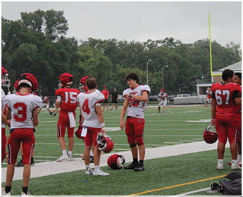 A group of people wearing red and white football uniforms stand on the sidelines of a football field with helmets in hand. Description generated by AI