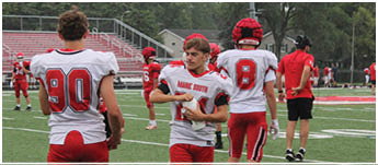 Three people in red and white football uniforms stand on a green field with others practicing in the background. Description generated by AI