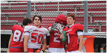 Three people wearing football uniforms stand in front of empty bleachers with a chain-link fence behind them. Description generated by AI