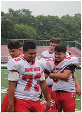 Four people wearing white and red football uniforms with “Maine South" on the front are standing together on a football field, with one person in the foreground smiling and others looking in different directions. Description generated by AI