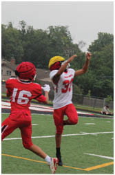 A person in red sports gear is preparing to throw a football on a field while another person in casual clothing stands near the sideline. Description generated by AI
