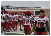 A group of people wearing red and white football uniforms are standing and walking on a football field near the end zone with a building and an American flag in the background. Description generated by AI