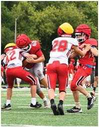 Children wearing helmets and football uniforms are actively engaged in a game on a grassy field with trees and houses in the background. Description generated by AI