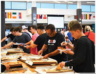 People stand in line indoors at a buffet or food-serving area with international flags displayed on the walls above. Description generated by AI