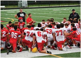 A group of people in red and white football uniforms kneel on a field while two adults stand nearby, one wearing a black shirt with text and the other in a black jacket and cap. Description generated by AI