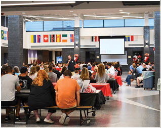 A large group of people seated at tables covered with red and black tablecloths attentively listening to a speaker in a spacious indoor area decorated with international flags. Description generated by AI