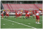 Children wearing red and white football uniforms and yellow helmets are playing a game on a green field with empty red bleachers in the background. Description generated by AI