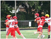 Children wearing helmets and football uniforms are playing a game on a grassy field with trees and a fence in the background. Description generated by AI