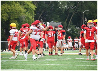 People wearing red and white football uniforms and helmets are gathered on a green field with trees in the background. Description generated by AI
