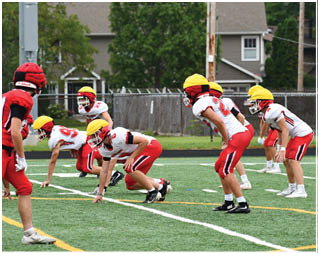 People wearing red and white football uniforms with yellow and red helmets are lined up on a green field preparing for a play. Description generated by AI