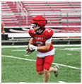 Two people in red football uniforms and helmets are playing on a green field with empty bleachers in the background. Description generated by AI