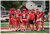 A group of people wearing red football uniforms and helmets are gathered on a field with a coach in a red shirt giving instructions. Description generated by AI