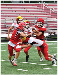 Four people in football uniforms are actively engaged in a tackle on a green field with empty bleachers in the background. Description generated by AI