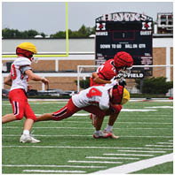 Three children wearing football uniforms and helmets are playing on a field with one child tackling another while a third child approaches. Description generated by AI