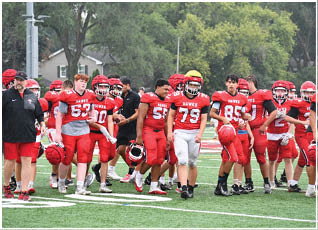 A group of people wearing red and white football uniforms and helmets are standing and walking on a green football field with trees and a building in the background. Description generated by AI
