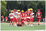 People wearing red and white football uniforms and yellow helmets are gathered on a green football field with trees in the background, with one person sitting on the ground. Description generated by AI