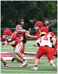 A group of people in red and white football uniforms are engaged in a play on a green field with a coach standing in the background. Description generated by AI