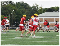 People wearing red and white football uniforms and yellow helmets are playing on a green football field with trees and a building in the background. Description generated by AI
