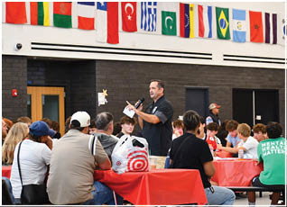 A person holding a microphone and paper is speaking to a group of people seated at tables covered with red tablecloths in a room decorated with various international flags. Description generated by AI