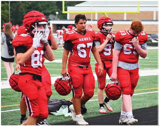 Four people in red football uniforms stand on a field, with two holding helmets and one adjusting a helmet. Description generated by AI