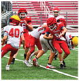 Children playing American football on a field with one player in red running with the ball while others in red and white uniforms attempt to tackle or block. Description generated by AI