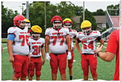 A group of five children in red and white football uniforms with yellow helmets stand on a football field while an adult in a red shirt and black cap gestures toward them. Description generated by AI