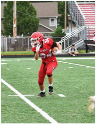 People in red and white football uniforms are playing on a green field with bleachers and trees in the background. Description generated by AI