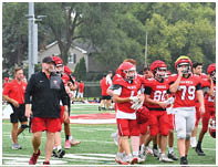 A group of people wearing red and white football uniforms and helmets are walking on a football field with a coach in black and red athletic wear leading them. Description generated by AI