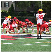 People wearing red and white football uniforms with yellow helmets are lined up on a football field preparing for a play. Description generated by AI