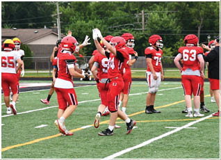 People wearing red and white football uniforms are gathered on a green field with trees and houses in the background. Description generated by AI
