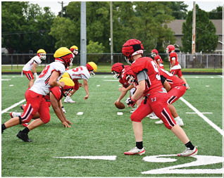 Two teams of people in football uniforms are lined up on a field facing each other, with one player holding the ball ready to snap it. Description generated by AI