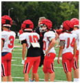 A group of people wearing red helmets and red and white football uniforms stand on a green field with one person in the background wearing black. Description generated by AI