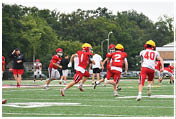 Children wearing red and white football uniforms with yellow helmets are playing on a green field with trees and a building in the background. Description generated by AI
