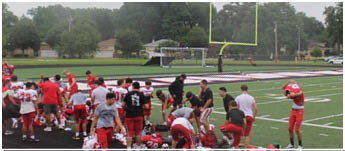A group of people in sports uniforms are gathered on the sidelines of a football field, some standing and some sitting or kneeling on the ground. Description generated by AI
