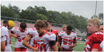 A group of people wearing red and white football uniforms are gathered on a field with trees and bleachers in the background. Description generated by AI