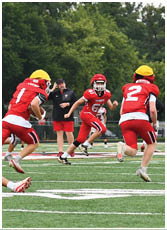 People in red football uniforms with yellow helmets are actively playing on a green field with trees in the background. Description generated by AI