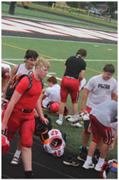 Three people in athletic clothing are on a sports field near a goalpost, with one person wearing a backpack and another bending down. Description generated by AI