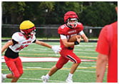 A person in red football gear with a helmet is running with the ball while another person in white and red gear with a yellow helmet reaches out to try to stop them on a football field. Description generated by AI