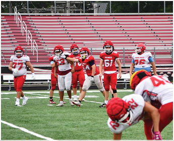 A group of people wearing red and white sports uniforms are kneeling and sitting together on a football field near the sideline. Description generated by AI